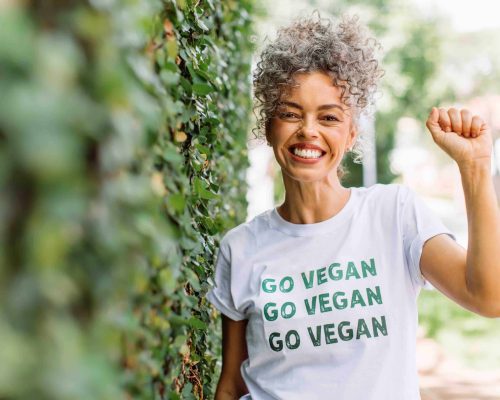 Vegan activist smiling cheerfully while standing alone outdoors. Happy mature woman advocating for veganism while wearing a shirt with the words "GO VEGAN" written on it.