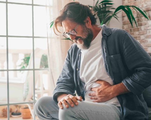 Bearded middle aged male in casual clothes touching belly and wincing in pain while sitting on sofa near window in living room at home